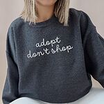 Woman in a Dark Heather crewneck sweatshirt embroidered with adopt don't shop in a light grey cursive script