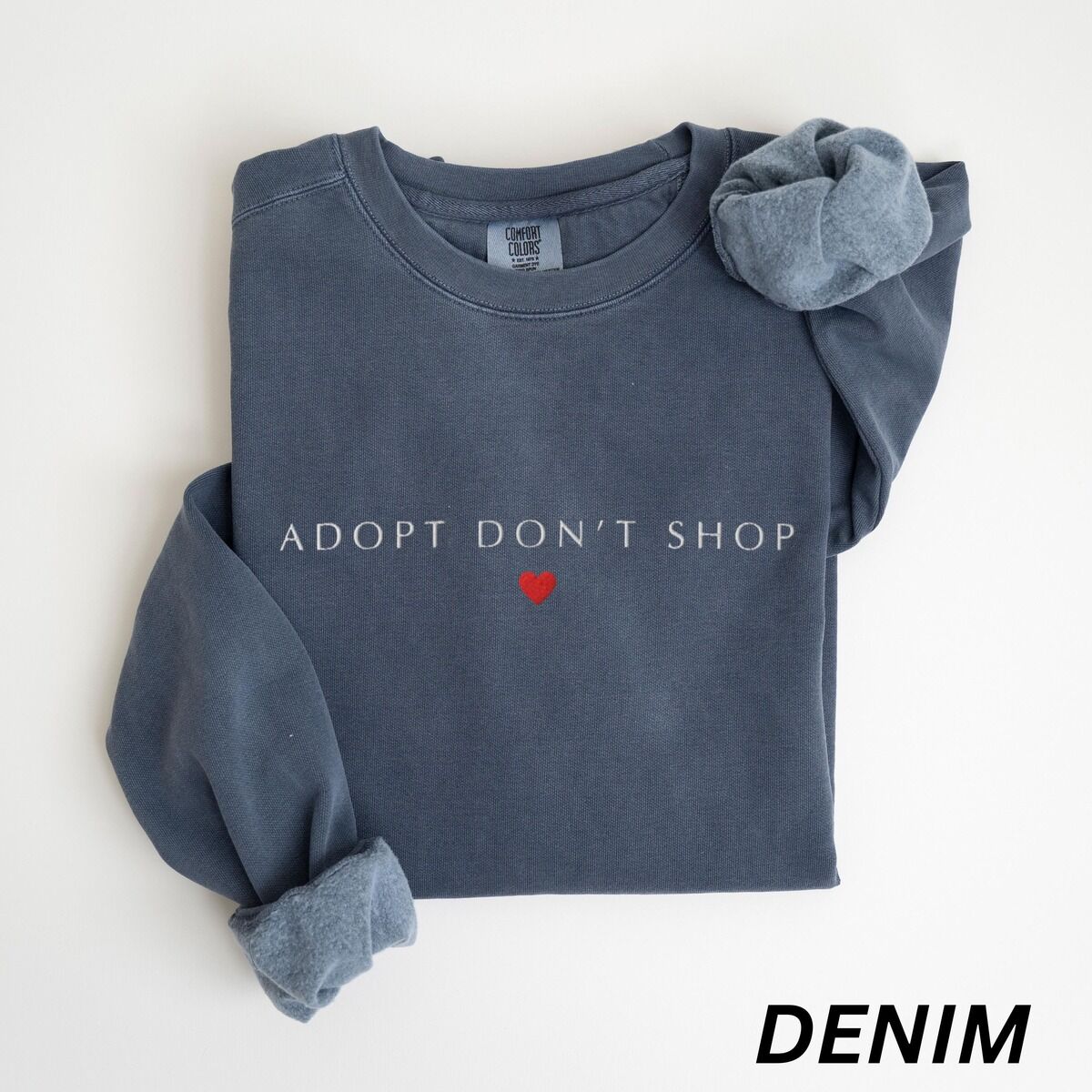 Adopt Don't Shop Sweatshirt – Denim Denim adopt don't shop sweatshirt laid flat on white with rolled cuffs, embroidered with ADOPT DON'T SHOP in white spaced capitals and a small red heart