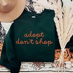 Forest Green crewneck sweatshirt laid flat with a hat and dried pampas, embroidered with adopt don't shop in a rust cursive script