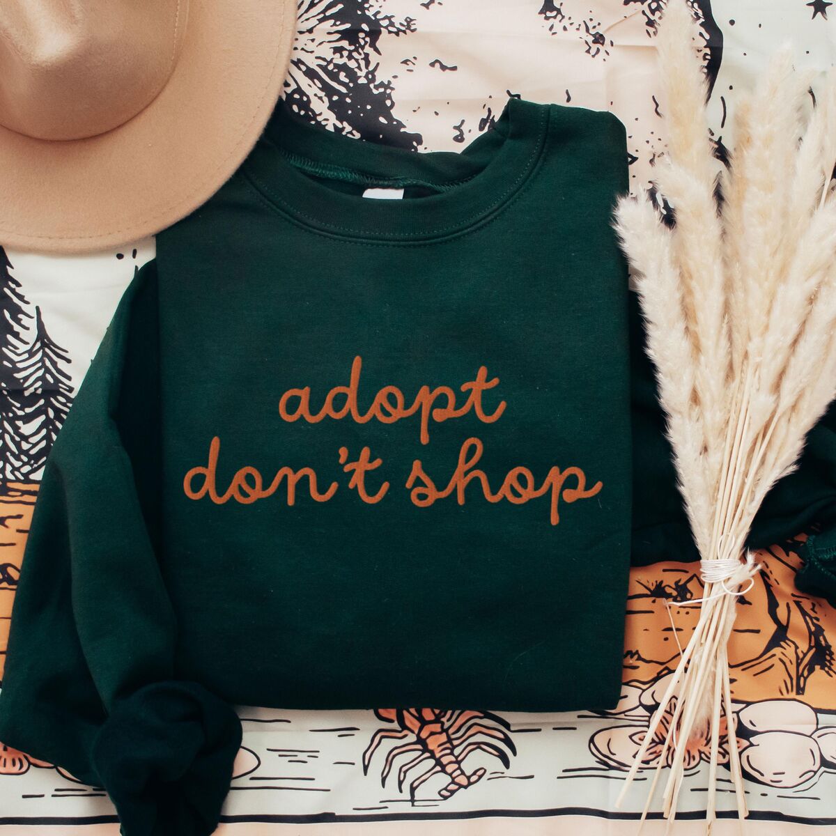 Adopt Don't Shop Sweatshirt - Forest Green Forest Green crewneck sweatshirt laid flat with a hat and dried pampas, embroidered with adopt don't shop in a rust cursive script