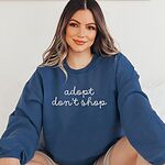 Smiling woman in an Indigo Blue crewneck sweatshirt embroidered with adopt don't shop in a light grey cursive script