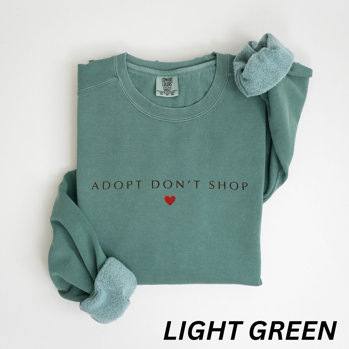 Adopt Don't Shop Sweatshirt – Light Green Light Green crewneck sweatshirt laid flat on white, embroidered with ADOPT DON'T SHOP in dark spaced capitals and a red heart