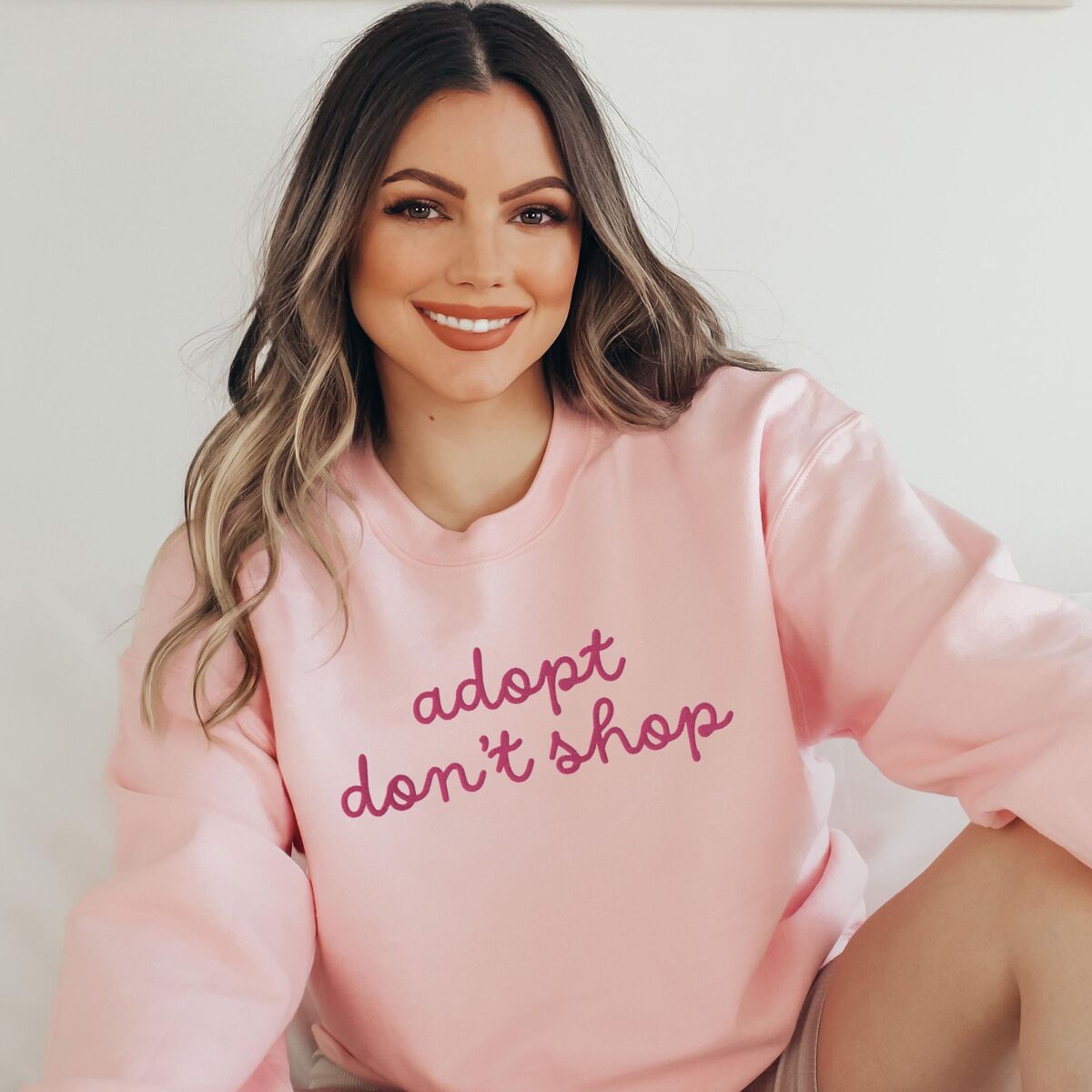 Adopt Don't Shop Sweatshirt - Light Pink Smiling woman in a Light Pink crewneck sweatshirt embroidered with adopt don't shop in a pink cursive script
