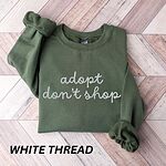 Military Green crewneck sweatshirt laid flat on wood, embroidered with adopt don't shop in a white cursive script on two lines