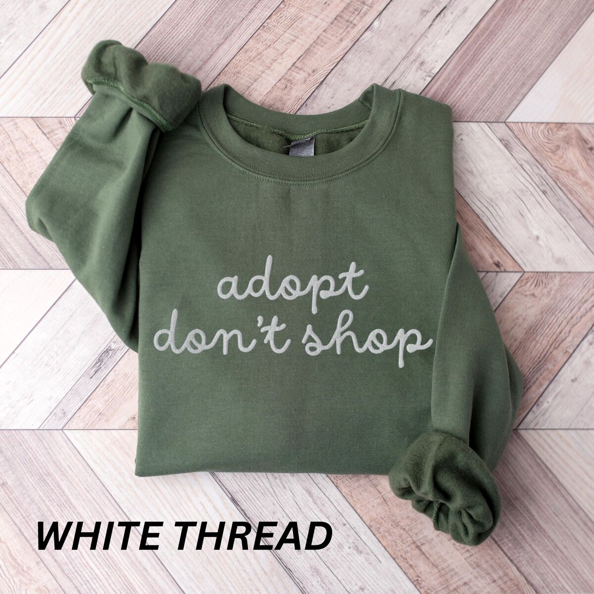 Adopt Don't Shop Sweatshirt - Military Green Military Green crewneck sweatshirt laid flat on wood, embroidered with adopt don't shop in a white cursive script on two lines