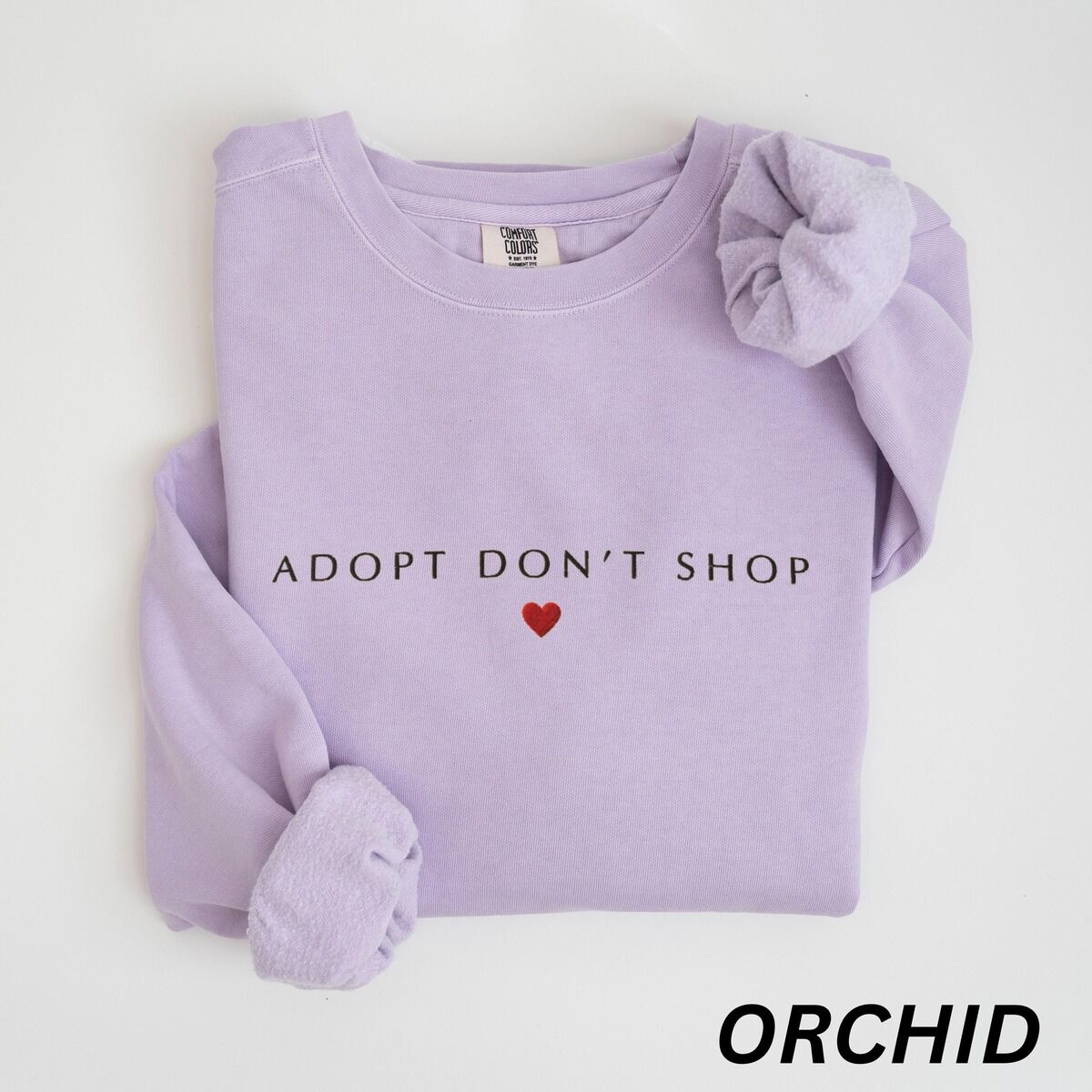 Adopt Don't Shop Sweatshirt – Orchid Orchid crewneck sweatshirt laid flat on white, embroidered with ADOPT DON'T SHOP in dark capitals and a small red heart