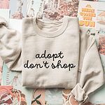 Sand crewneck sweatshirt laid flat on a collage of postcards, embroidered with adopt don't shop in a black cursive script