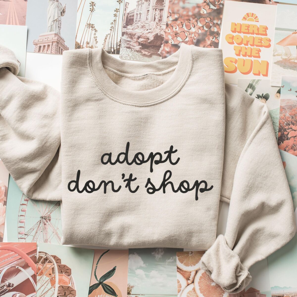 Adopt Don't Shop Sweatshirt - Sand Sand crewneck sweatshirt laid flat on a collage of postcards, embroidered with adopt don't shop in a black cursive script