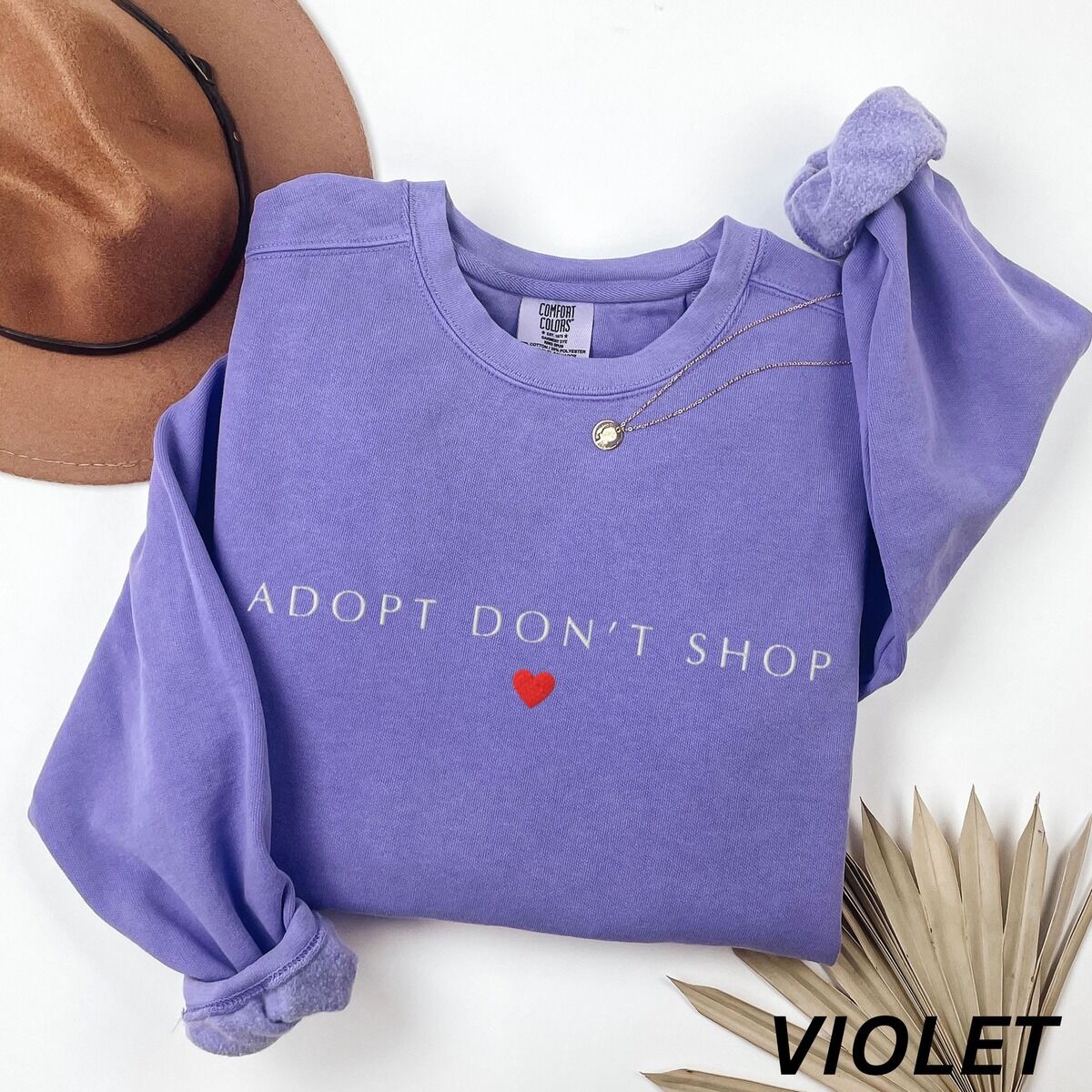 Adopt Don't Shop Sweatshirt – Violet Violet crewneck sweatshirt laid flat with a felt hat and dried palm leaves, embroidered with ADOPT DON'T SHOP in light capitals and a red heart