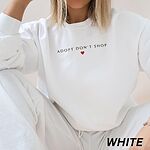 Woman sitting in a White crewneck sweatshirt embroidered with ADOPT DON'T SHOP in dark capitals and a small red heart