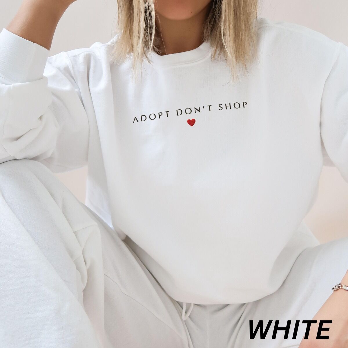 Adopt Don't Shop Sweatshirt – White Woman sitting in a White crewneck sweatshirt embroidered with ADOPT DON'T SHOP in dark capitals and a small red heart