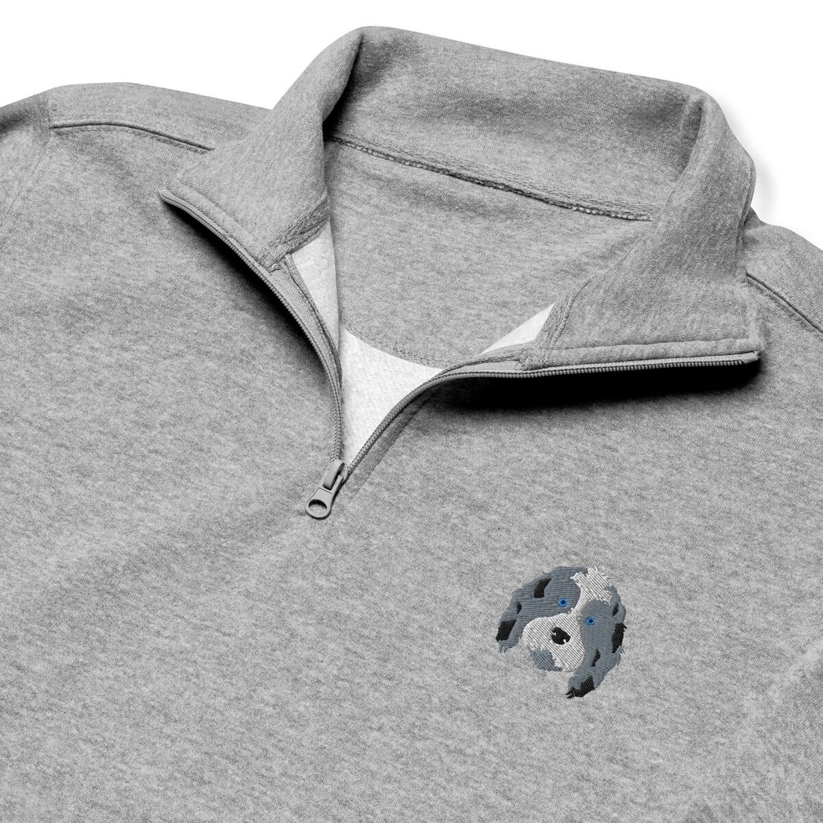 Aussiedoodle Quarter Zip - close-up Close-up of a heather grey quarter zip collar and chest, embroidered with a blue merle aussiedoodle face with a white blaze and blue eyes on the left chest