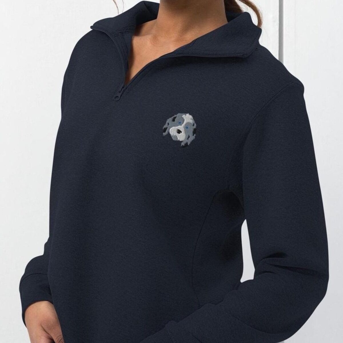 Aussiedoodle Quarter Zip - worn, side view Woman in a Navy quarter zip pullover with the collar open, embroidered with a blue merle aussiedoodle face with a white blaze and blue eyes on the left chest
