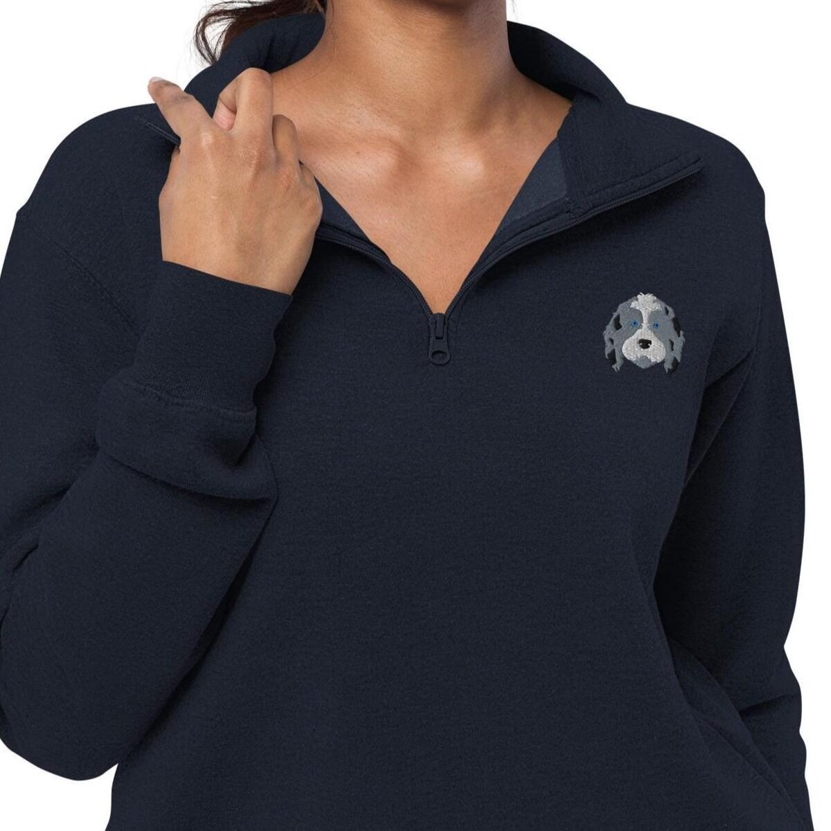 Aussiedoodle Quarter Zip - worn Woman holding the zip collar of a Navy quarter zip pullover embroidered with a blue merle aussiedoodle face with a white blaze and blue eyes on the left chest