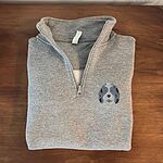 Folded Sport Grey quarter zip pullover on a wooden table, embroidered with a blue merle aussiedoodle face with a white blaze and blue eyes on the left chest