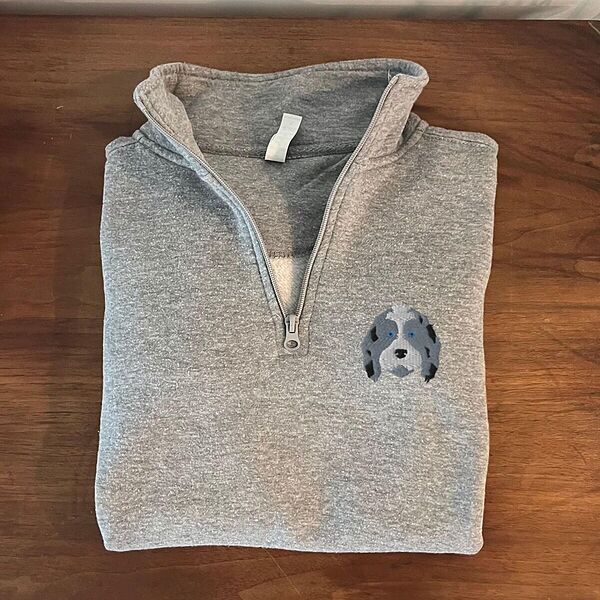 Folded Sport Grey quarter zip pullover on a wooden table, embroidered with a blue merle aussiedoodle face with a white blaze and blue eyes on the left chest