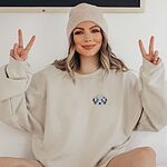 Woman in a beanie making peace signs in a Sand crewneck sweatshirt embroidered with a fluffy grey aussiedoodle face with spotted merle ears and blue eyes on the left chest