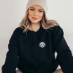 Woman in a beanie wearing a Black crewneck sweatshirt embroidered with a fluffy grey aussiedoodle face with spotted merle ears and blue eyes on the left chest