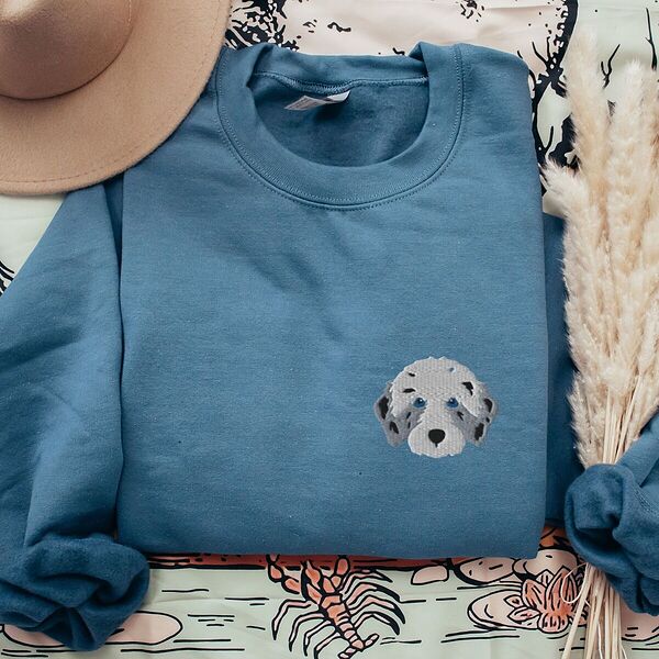Indigo Blue crewneck sweatshirt laid flat with a tan hat and dried pampas grass, embroidered with a fluffy grey aussiedoodle face with spotted merle ears and blue eyes on the left chest