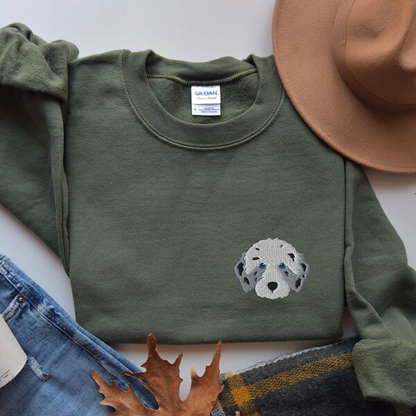 Military Green crewneck sweatshirt laid flat with a tan hat, jeans, a plaid scarf and autumn leaves, embroidered with a fluffy grey aussiedoodle face with spotted merle ears and blue eyes on the left chest