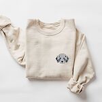 Sand crewneck sweatshirt laid flat on a white background, embroidered with a fluffy grey aussiedoodle face with spotted merle ears and blue eyes on the left chest