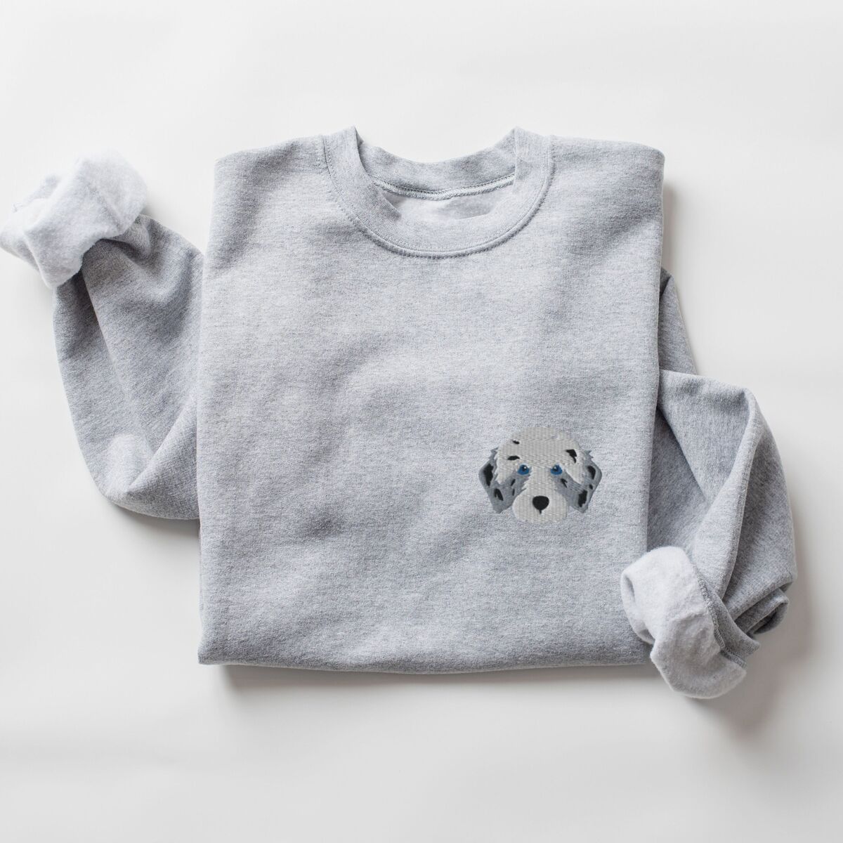Aussiedoodle Sweatshirt - Sport Grey Sport Grey crewneck sweatshirt laid flat on a white background, embroidered with a fluffy grey aussiedoodle face with spotted merle ears and blue eyes on the left chest