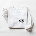 White crewneck sweatshirt laid flat on a white background, embroidered with a fluffy grey aussiedoodle face with spotted merle ears and blue eyes on the left chest