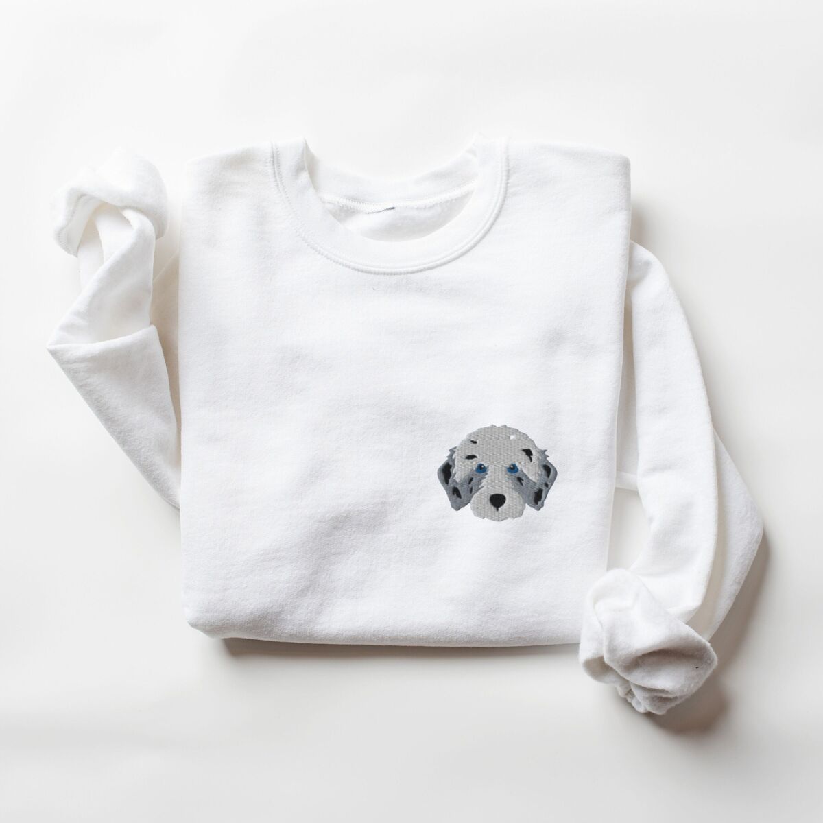 Aussiedoodle Sweatshirt - White White crewneck sweatshirt laid flat on a white background, embroidered with a fluffy grey aussiedoodle face with spotted merle ears and blue eyes on the left chest