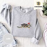 Ash crewneck sweatshirt folded with denim shorts, white sneakers and a potted plant, embroidered with a blue merle Australian Shepherd in a Santa hat trotting with a string of Christmas lights in its mouth