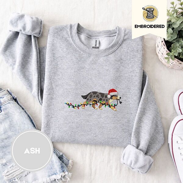 Ash crewneck sweatshirt folded with denim shorts, white sneakers and a potted plant, embroidered with a blue merle Australian Shepherd in a Santa hat trotting with a string of Christmas lights in its mouth