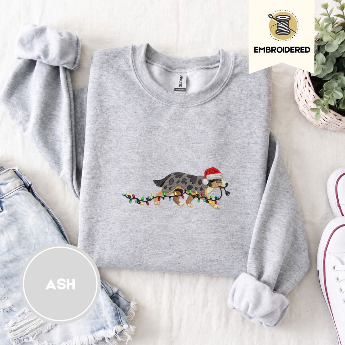 Australian Shepherd Christmas Sweatshirt – Ash Ash crewneck sweatshirt folded with denim shorts, white sneakers and a potted plant, embroidered with a blue merle Australian Shepherd in a Santa hat trotting with a string of Christmas lights in its mouth
