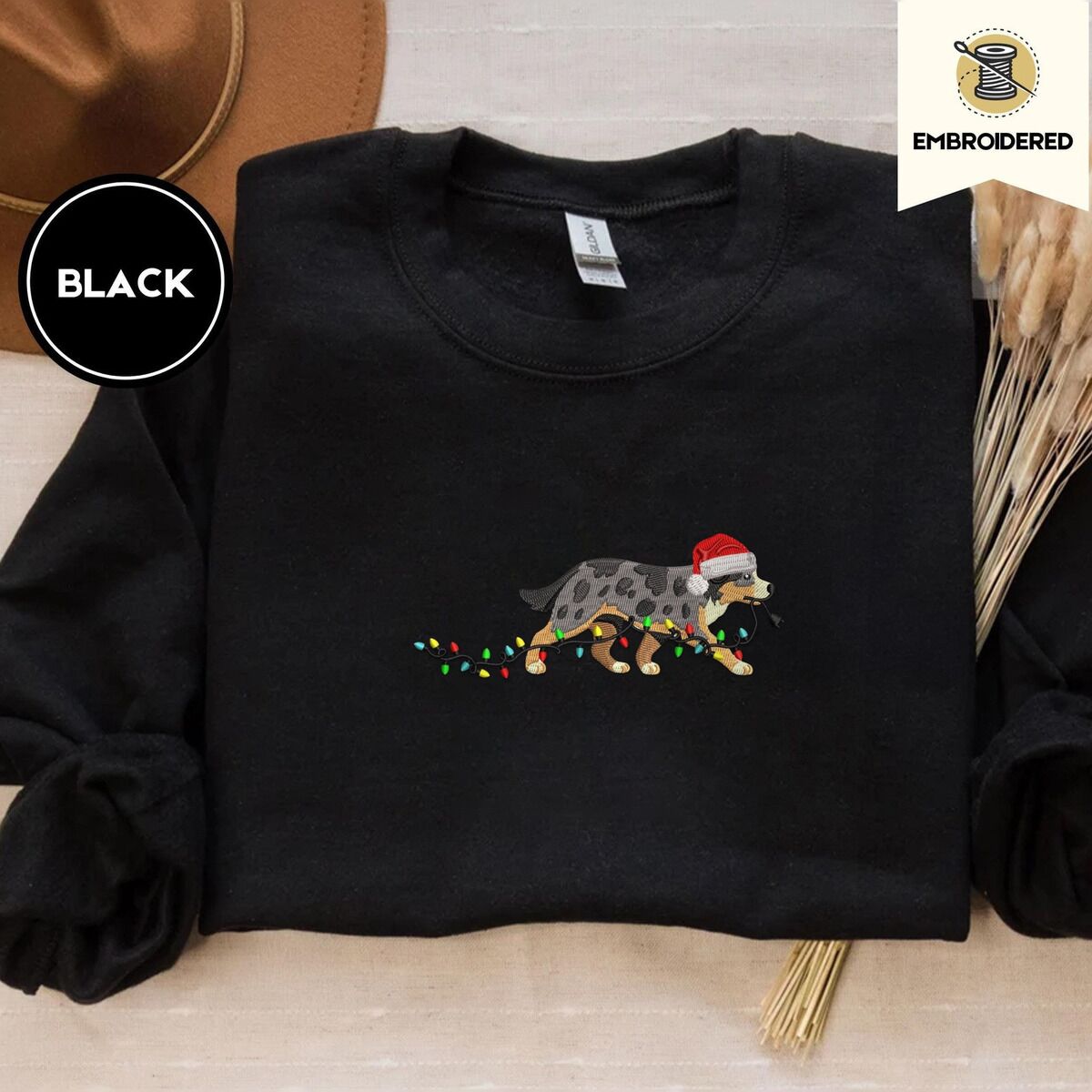Australian Shepherd Christmas Sweatshirt – Black Black crewneck sweatshirt with a brown felt hat and dried bunny tail grass, embroidered with a blue merle Australian Shepherd in a Santa hat trotting with a string of Christmas lights in its mouth