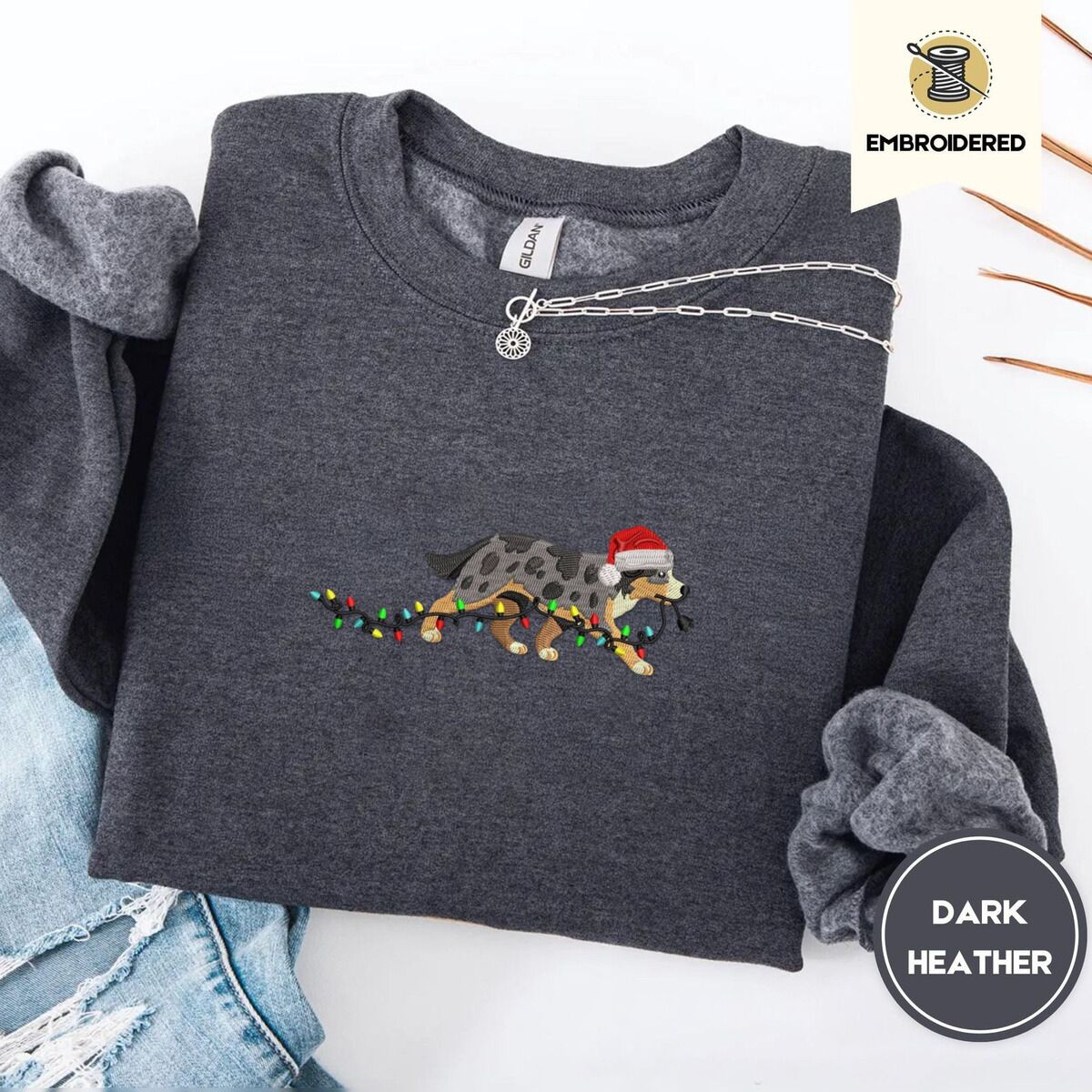 Australian Shepherd Christmas Sweatshirt – Dark Heather Dark Heather crewneck sweatshirt folded with ripped jeans and a silver chain necklace, embroidered with a blue merle Australian Shepherd in a Santa hat trotting with a string of Christmas lights in its mouth