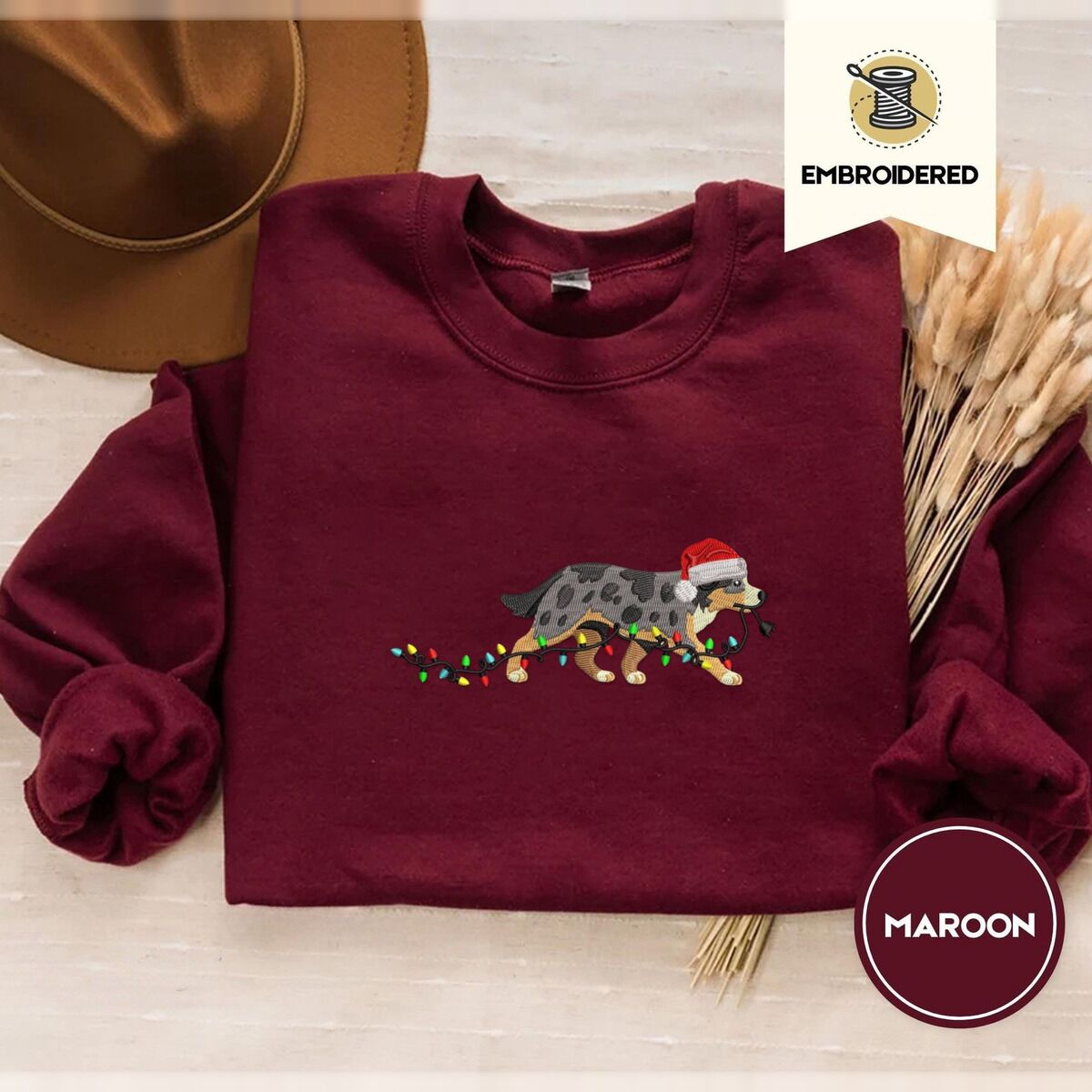 Australian Shepherd Christmas Sweatshirt – Maroon Maroon crewneck sweatshirt with a brown felt hat and dried bunny tail grass, embroidered with a blue merle Australian Shepherd in a Santa hat trotting with a string of Christmas lights in its mouth