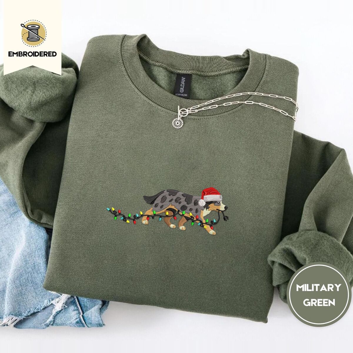 Australian Shepherd Christmas Sweatshirt – Military Green Military Green crewneck sweatshirt folded with light jeans and a silver chain necklace, embroidered with a blue merle Australian Shepherd in a Santa hat trotting with a string of Christmas lights in its mouth