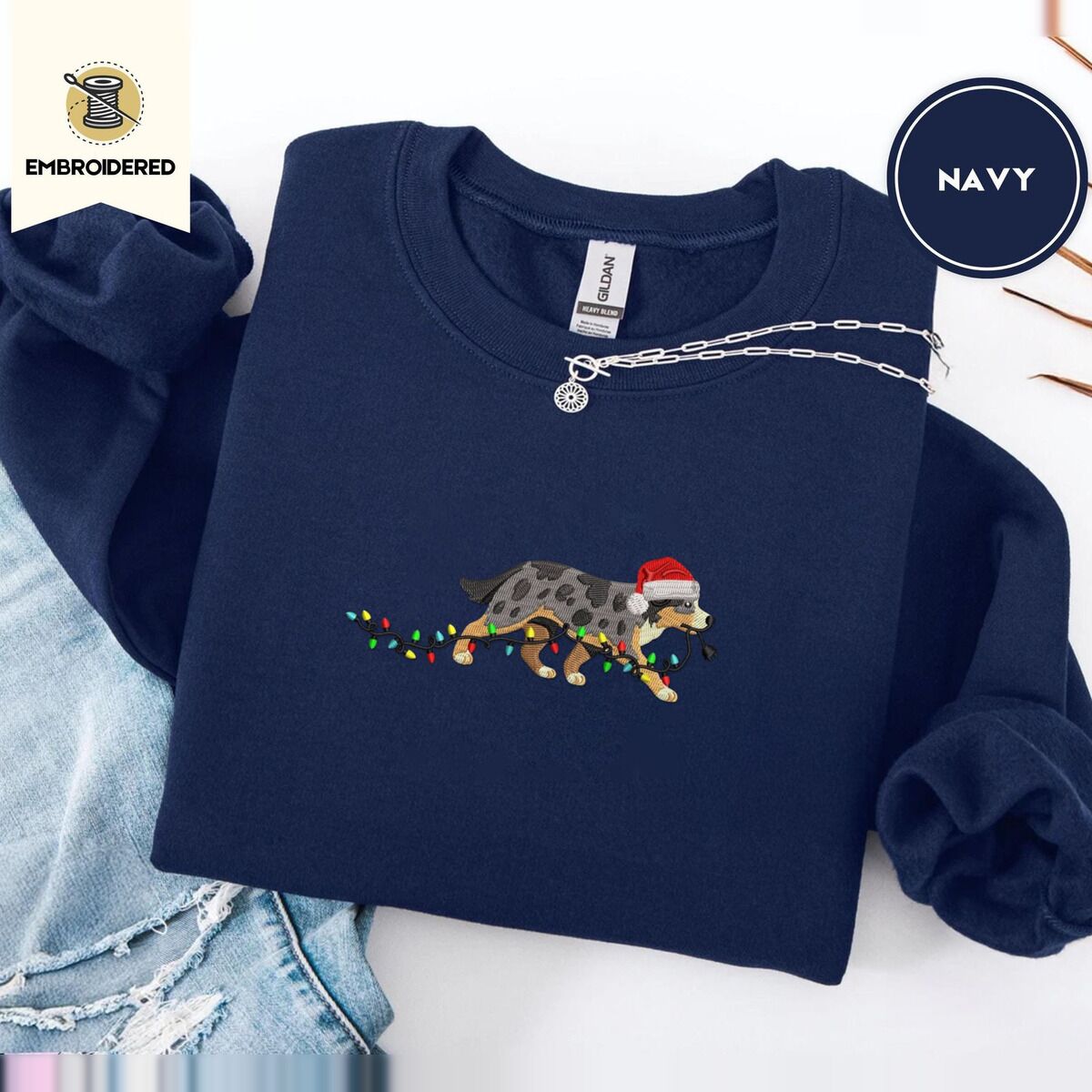 Australian Shepherd Christmas Sweatshirt – Navy Navy crewneck sweatshirt folded with ripped jeans and a silver chain necklace, embroidered with a blue merle Australian Shepherd in a Santa hat trotting with a string of Christmas lights in its mouth