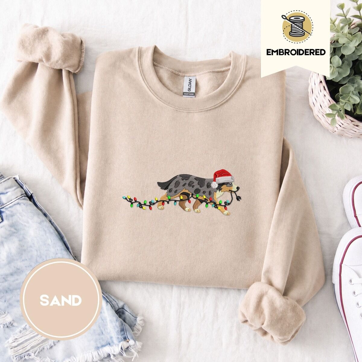 Australian Shepherd Christmas Sweatshirt – Sand Sand crewneck sweatshirt folded with denim shorts, white sneakers and a potted plant, embroidered with a blue merle Australian Shepherd in a Santa hat trotting with a string of Christmas lights in its mouth