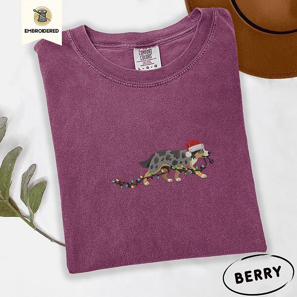 Berry Comfort Colors T-shirt embroidered with a blue merle Australian Shepherd in a Santa hat trotting with a string of Christmas lights in its mouth
