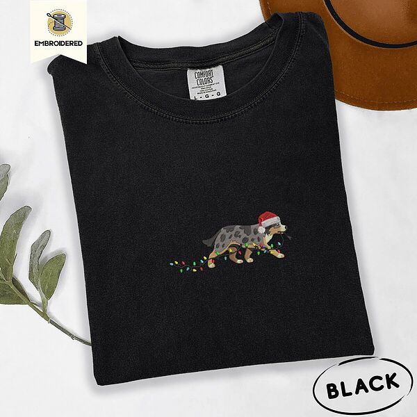 Black Comfort Colors T-shirt embroidered with a blue merle Australian Shepherd in a Santa hat trotting with a string of Christmas lights in its mouth
