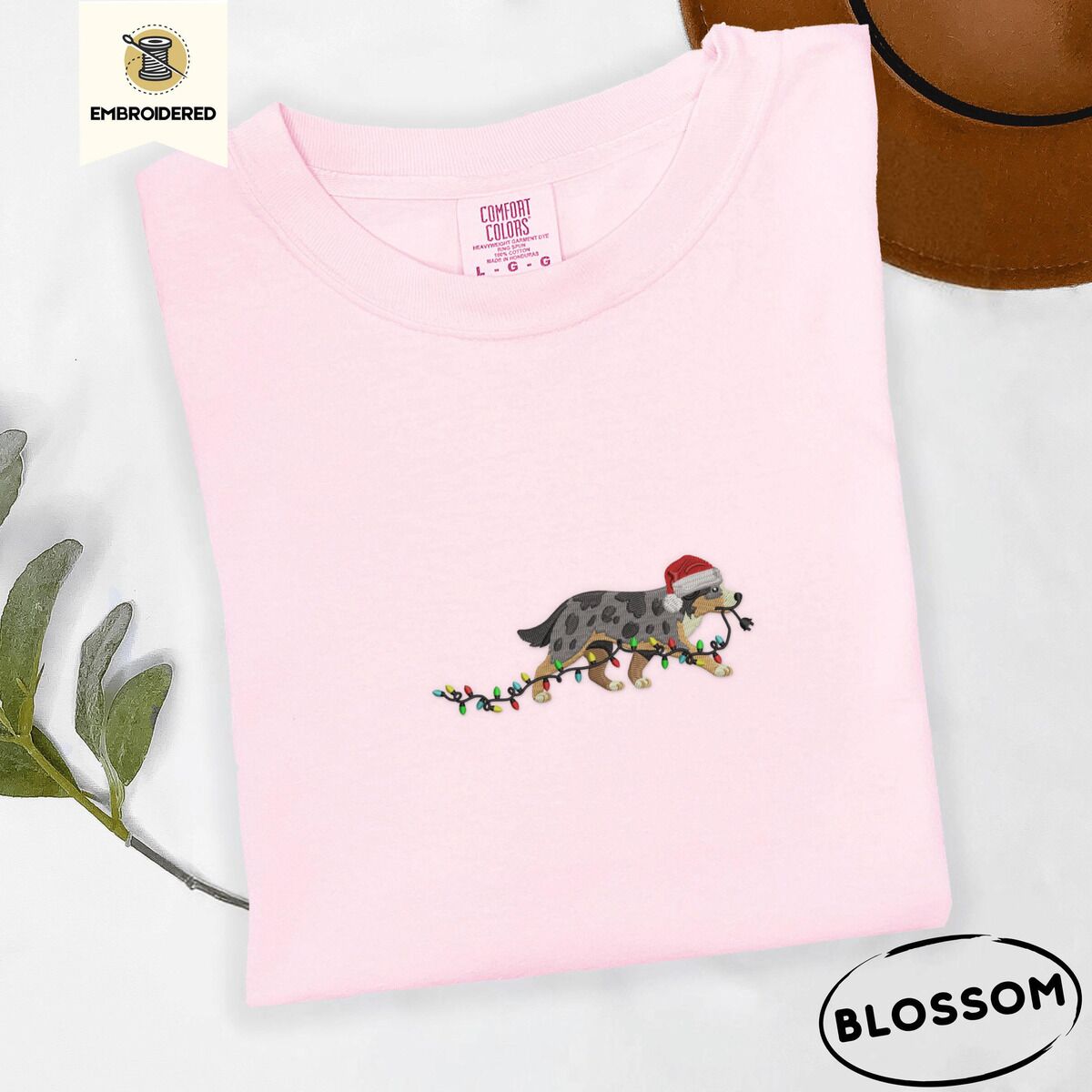 Australian Shepherd Santa Hat Shirt – Blossom Blossom Comfort Colors T-shirt embroidered with a blue merle Australian Shepherd in a Santa hat trotting with a string of Christmas lights in its mouth