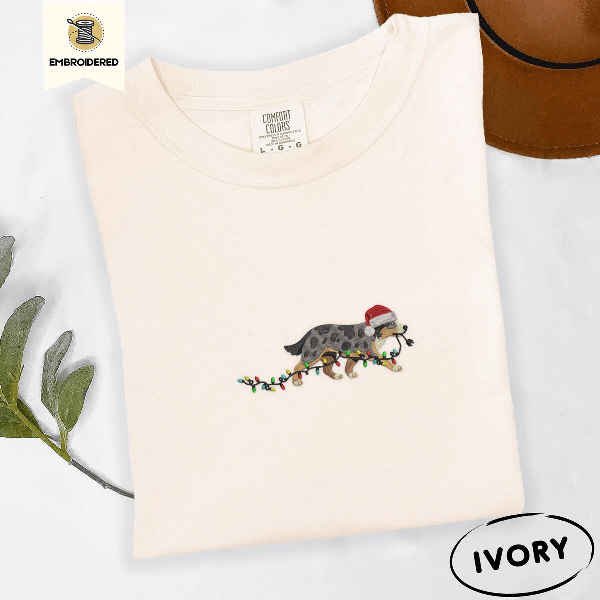 Australian Shepherd Santa Hat Shirt – Ivory Ivory Comfort Colors T-shirt embroidered with a blue merle Australian Shepherd in a Santa hat trotting with a string of Christmas lights in its mouth