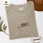 Khaki Comfort Colors T-shirt embroidered with a blue merle Australian Shepherd in a Santa hat trotting with a string of Christmas lights in its mouth