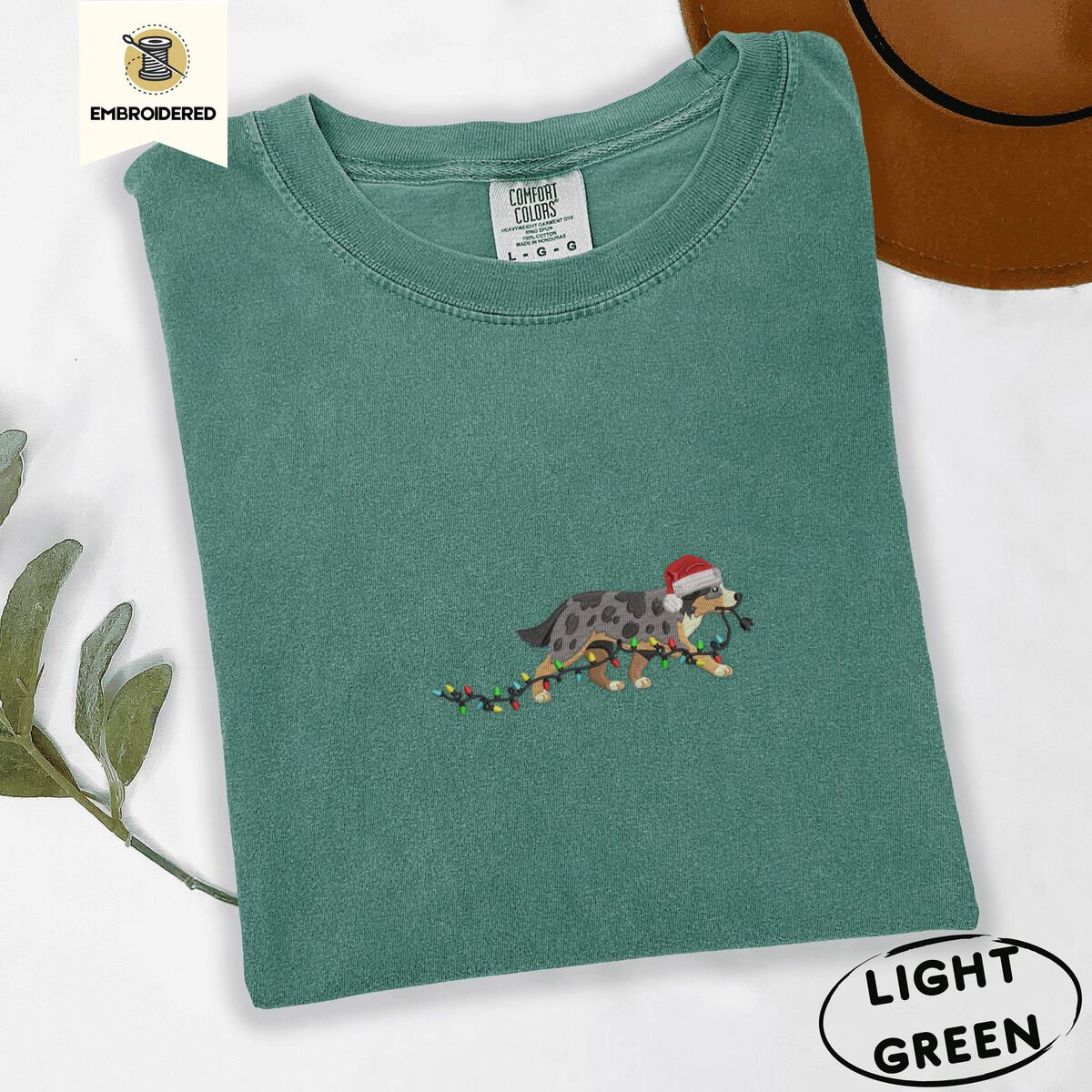 Australian Shepherd Santa Hat Shirt – Light Green Light Green Comfort Colors T-shirt embroidered with a blue merle Australian Shepherd in a Santa hat trotting with a string of Christmas lights in its mouth