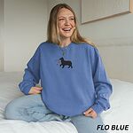 Smiling woman sitting on a bed in a bright blue crewneck sweatshirt embroidered with a black Australian shepherd silhouette carrying a string of Christmas lights
