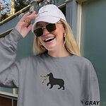 Smiling woman in a cap and sunglasses in a Grey crewneck sweatshirt embroidered with a black Australian shepherd silhouette carrying a string of Christmas lights