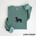 Light Green crewneck sweatshirt laid flat with rolled cuffs, embroidered with a black Australian shepherd silhouette carrying a string of Christmas lights
