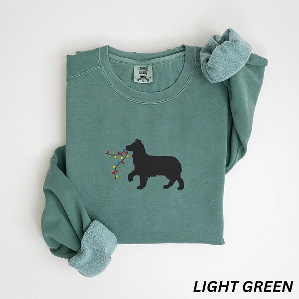 Light Green crewneck sweatshirt laid flat with rolled cuffs, embroidered with a black Australian shepherd silhouette carrying a string of Christmas lights