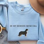 Light Blue crewneck sweatshirt laid flat with a coffee cup and a felt hat, embroidered with In My Berner Mom Era above a standing Bernese mountain dog