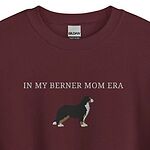 Close view of a Maroon crewneck sweatshirt embroidered with In My Berner Mom Era in light thread above a standing Bernese mountain dog