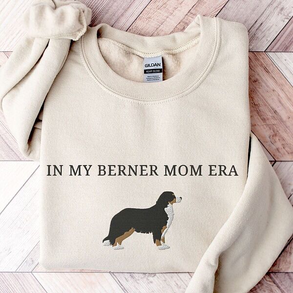 Sand Berner mom era sweatshirt laid flat on whitewashed wood, embroidered with In My Berner Mom Era above a standing tricolor Bernese mountain dog